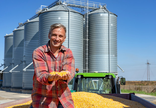 Happy,Farmer,Showing,Freshly,Harvested,Corn,Grains,Against,Grain,Silos.
